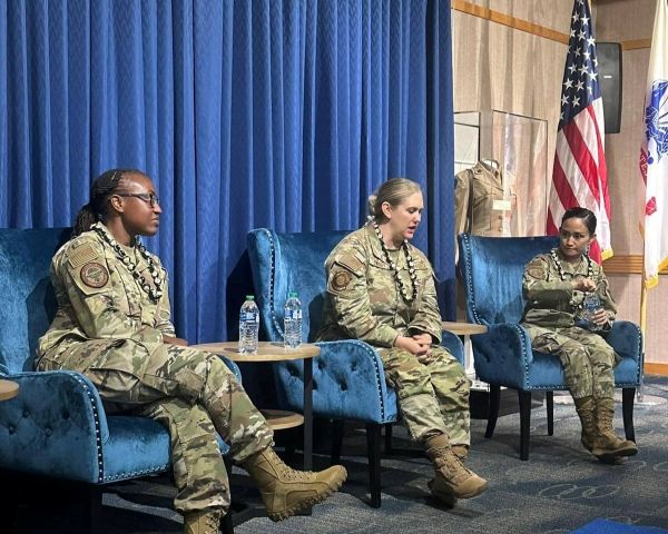 Photo of 3 women sitting on charis giving a briefing with the American flag in the background.